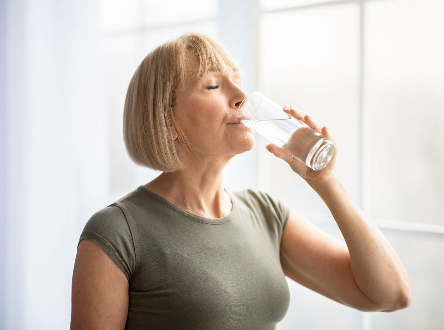 Older woman in an olive tee drinking a glass of water by a bright window