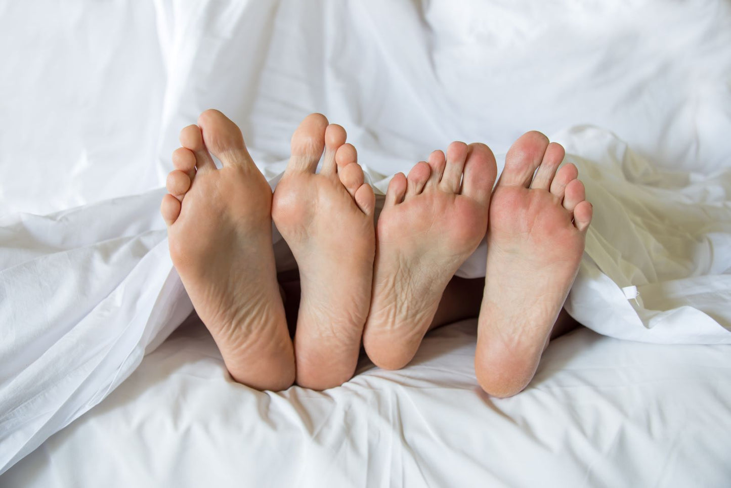 Two pairs of feet peeking from under white bed sheets, representing intimacy and sexual health for couples