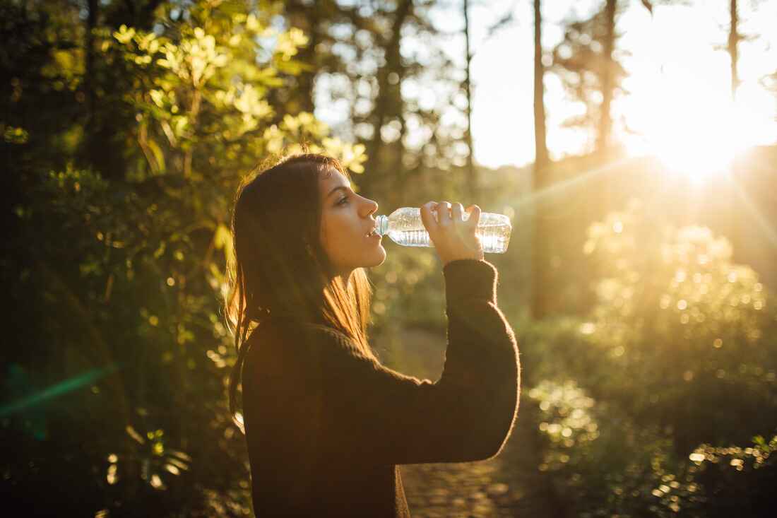 Woman drinking from a water bottle outdoors, backlit by the setting sun through trees