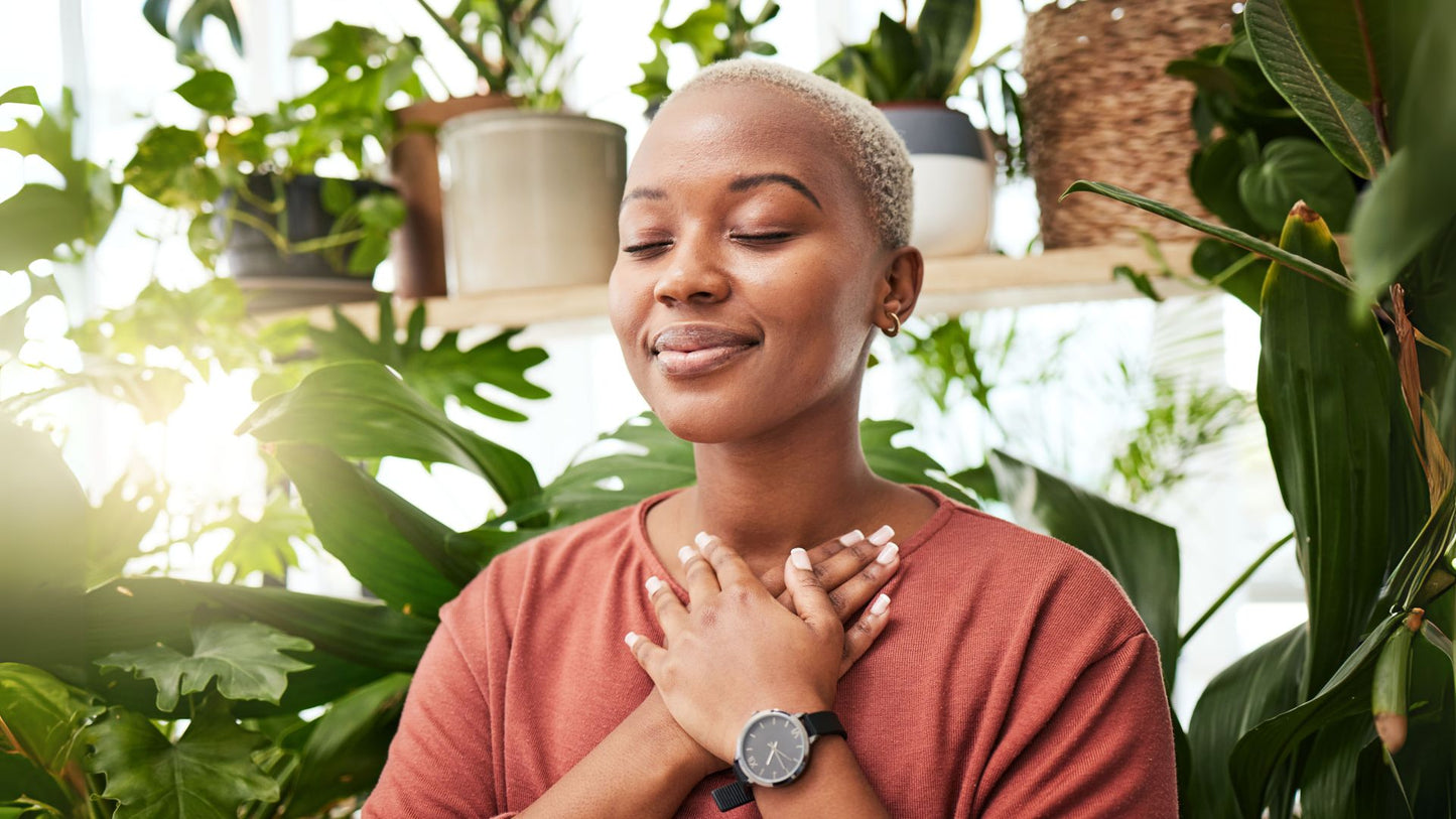 Smiling woman with hands over her heart, eyes closed, surrounded by houseplants