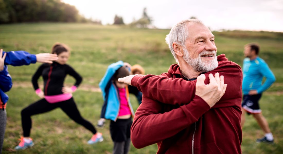 Older man with a white beard stretching his arm during an outdoor group fitness class on grass