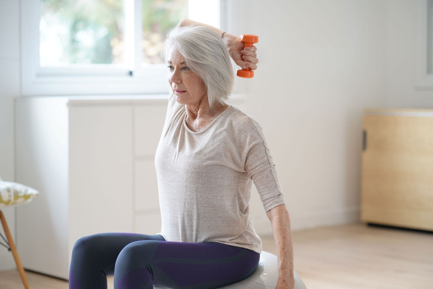 Silver-haired woman raising an orange dumbbell while seated on an exercise ball