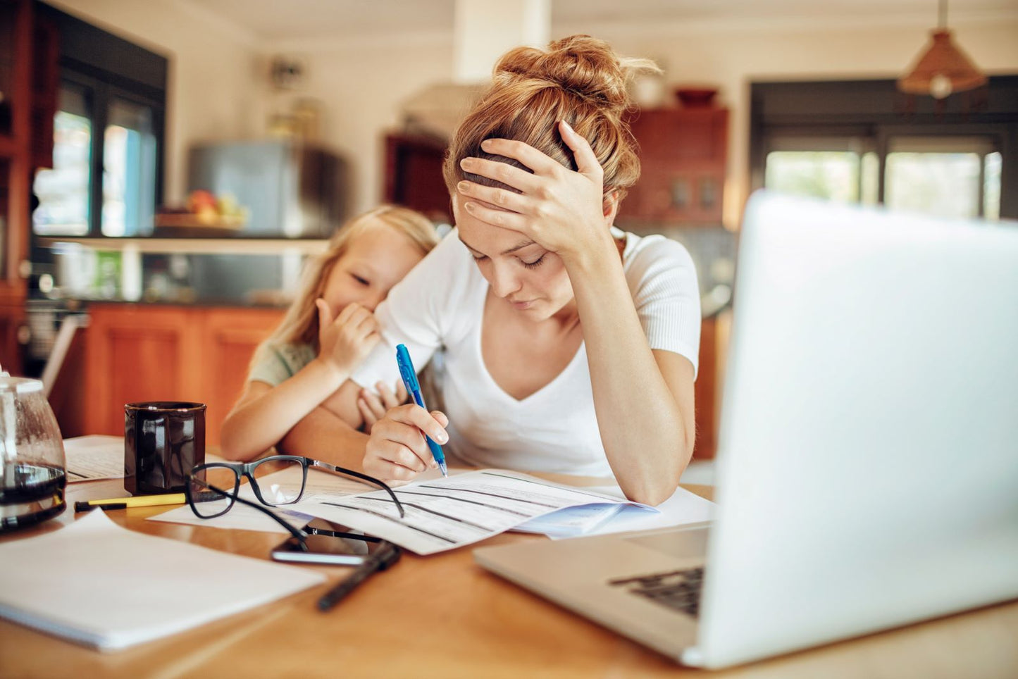 Stressed woman at a table with paperwork and a laptop, hand to her head, child beside her