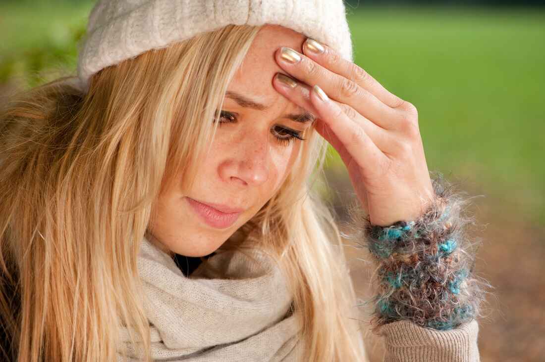 Woman in a knit hat and scarf outdoors holding her forehead, looking unwell in cold weather