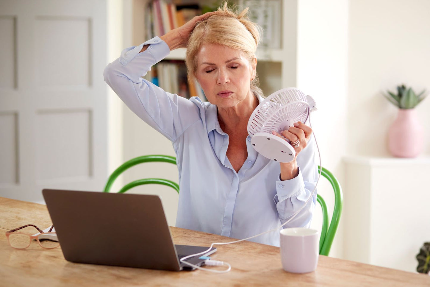 Older woman at a laptop fanning herself with a small fan during a hot flash