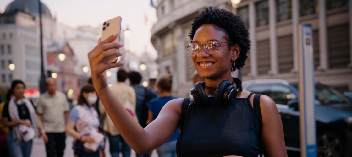 Smiling woman with glasses taking a photo on her phone on a busy city street