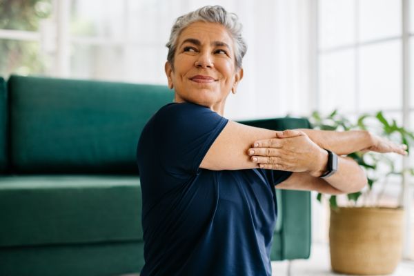 Older woman stretching her arm across her chest in a bright living room