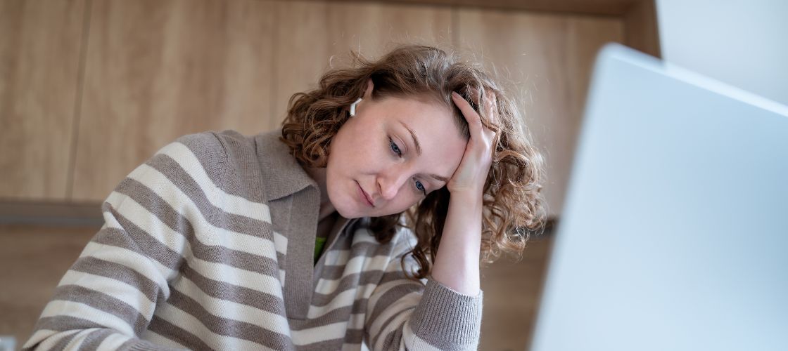 Tired woman in a striped sweater resting her head on her hand while looking at a laptop