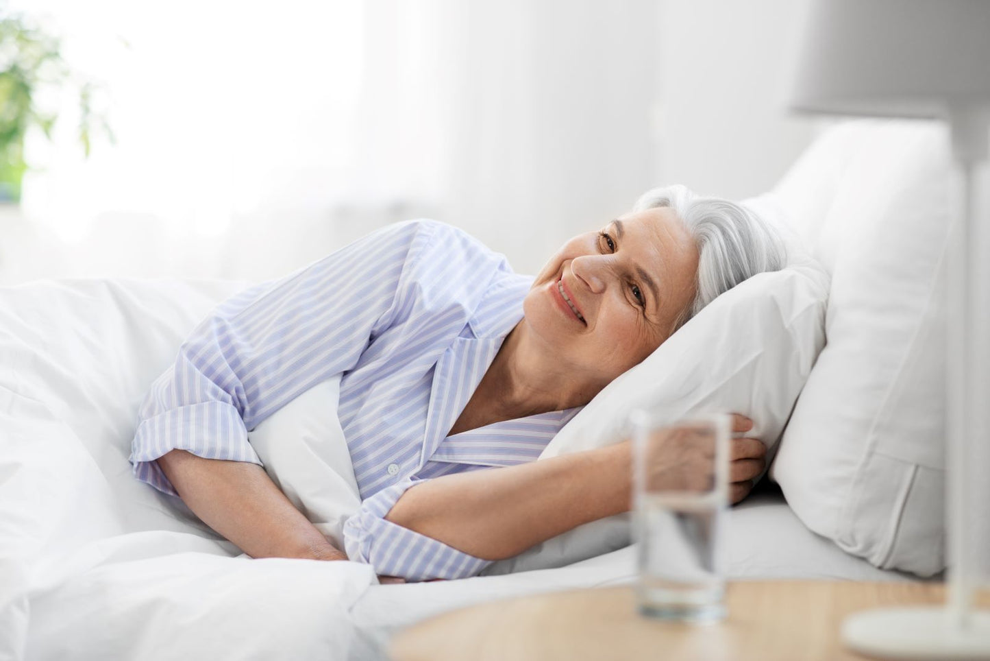 Smiling older woman lying in bed in striped pajamas with water on the nightstand