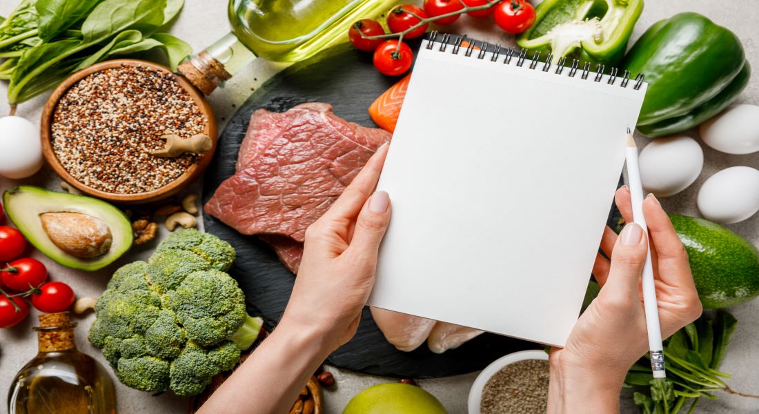 Hands with a notepad over a spread of keto foods—meat, salmon, avocado, and greens—for planning potassium intake