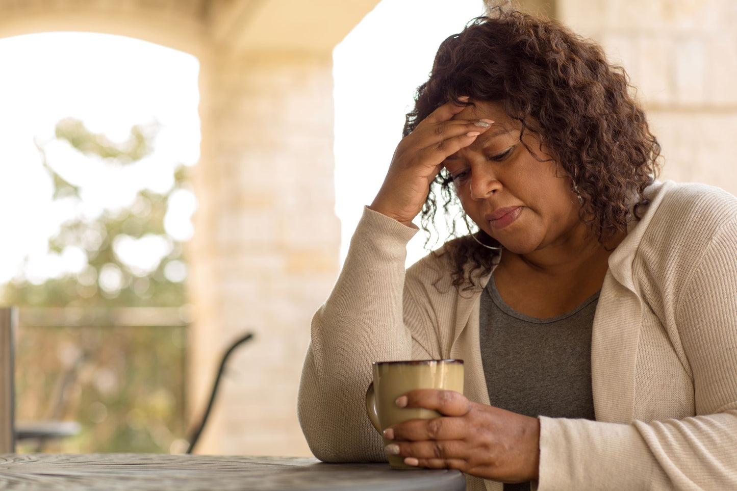 Woman resting her forehead in one hand while holding a mug at an outdoor table, looking tired