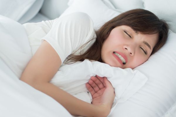 Woman asleep in bed grimacing and grinding her teeth, illustrating bruxism and TMJ tension