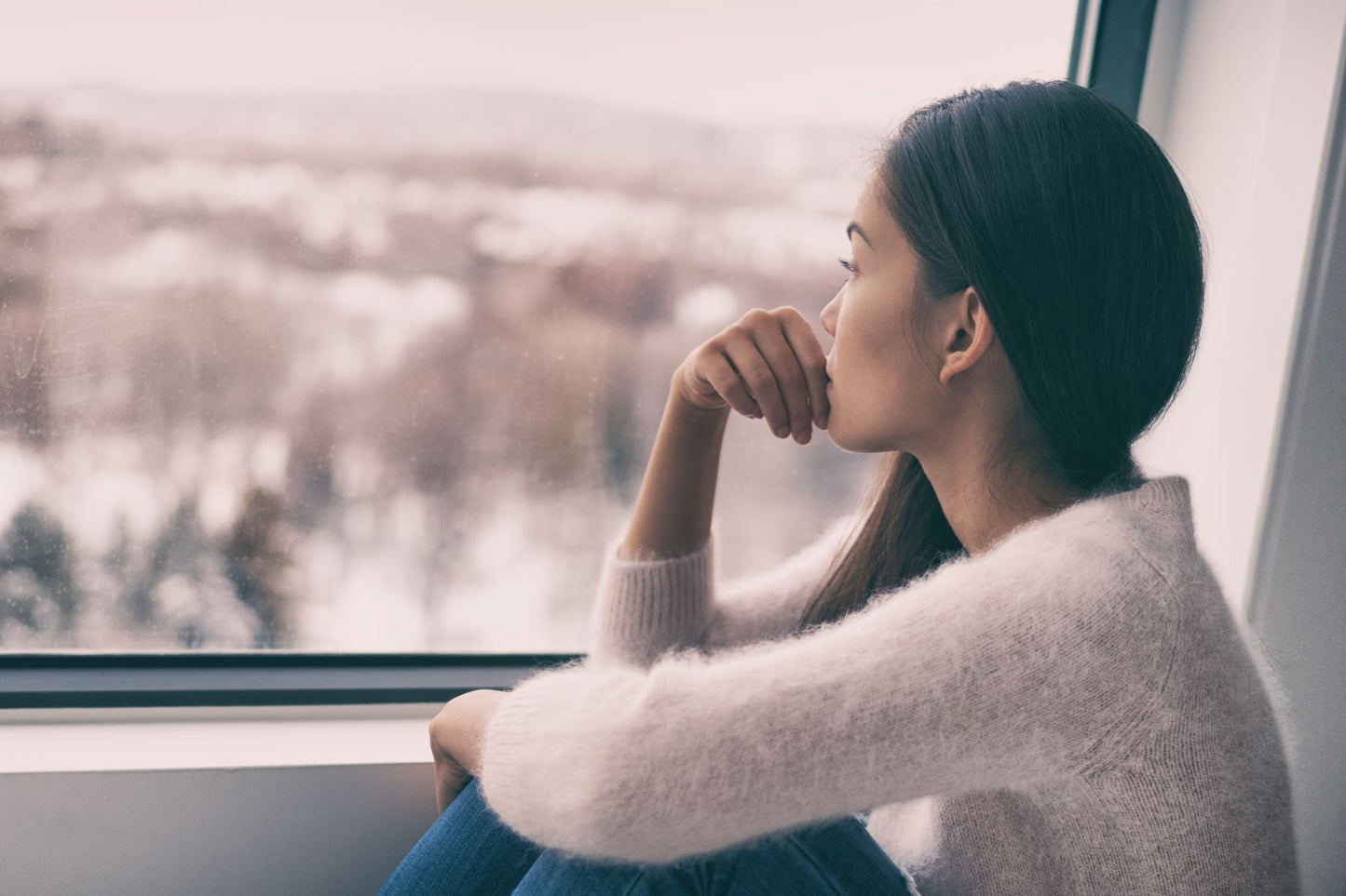 Woman in a sweater sitting by a window gazing out at a wintry landscape, looking pensive
