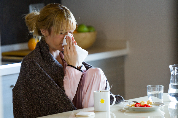 Woman wrapped in a blanket blowing her nose with a tissue at a table