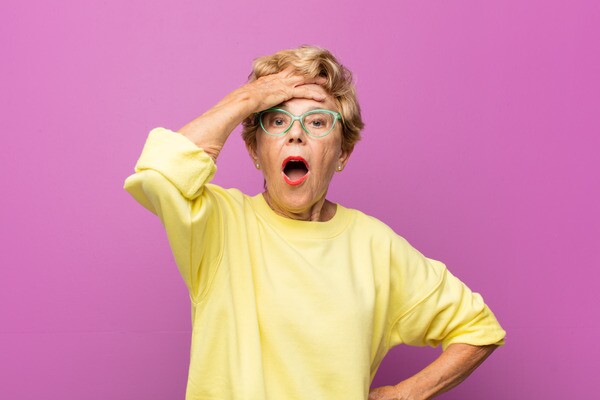 Surprised older woman in a yellow sweater on a pink background, hand to her forehead