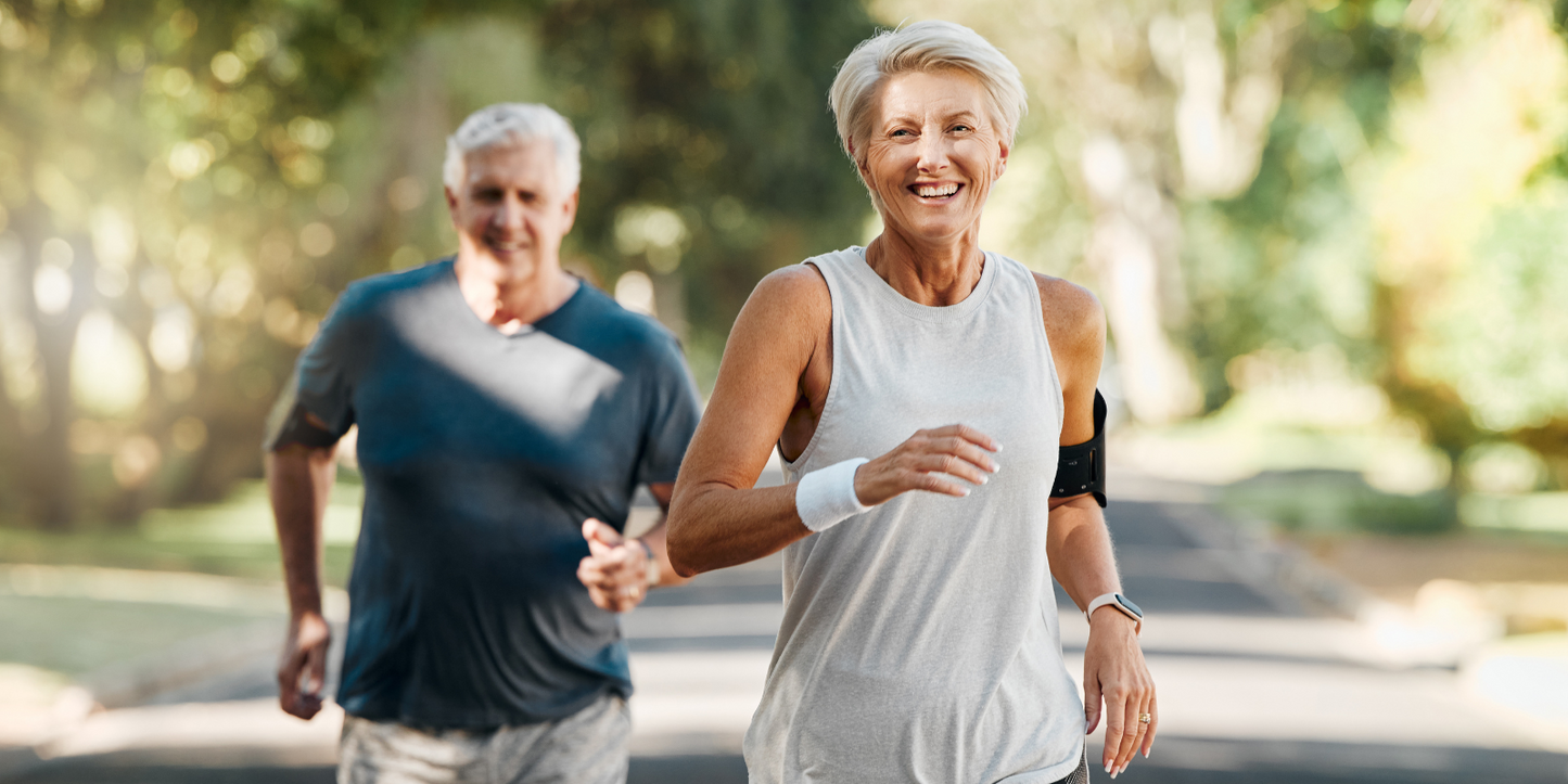Older couple jogging outdoors on a tree-lined path, smiling woman in front
