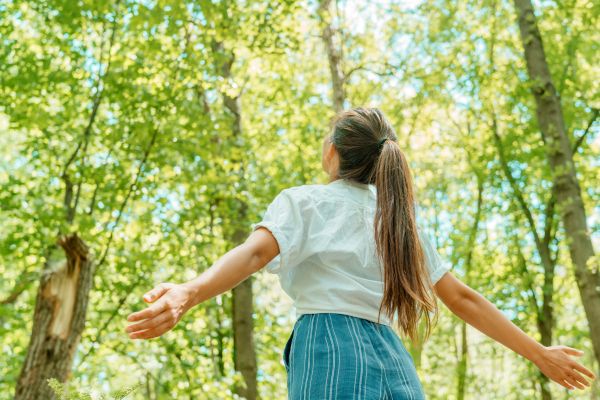 Woman in a white blouse standing with arms outstretched in a sunlit green forest