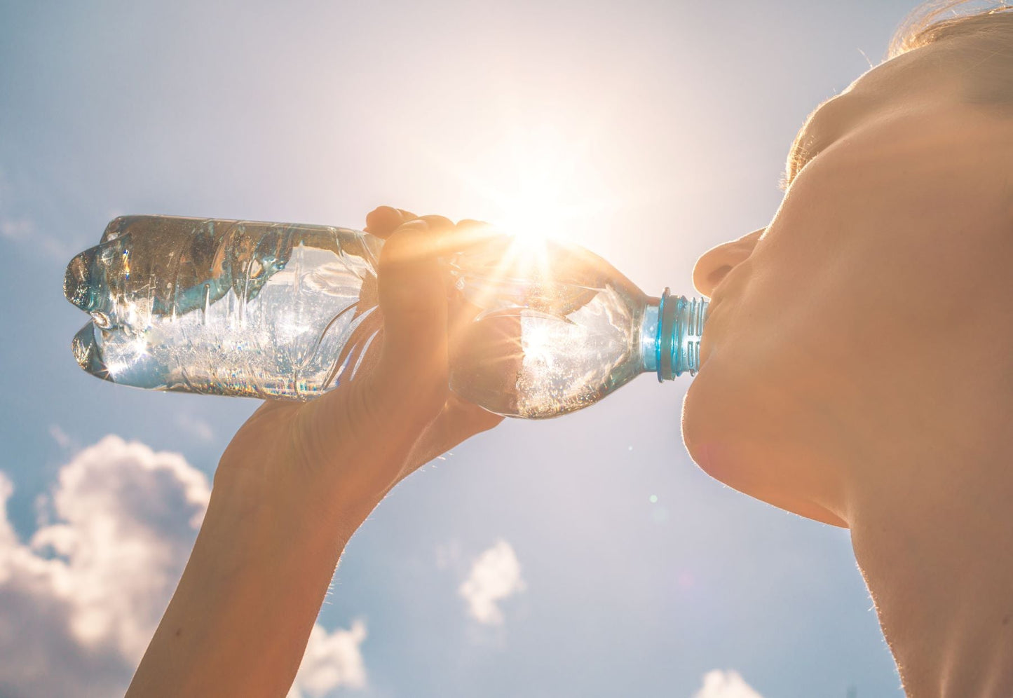 Person drinking from a clear water bottle against a bright sunlit sky, illustrating hydration